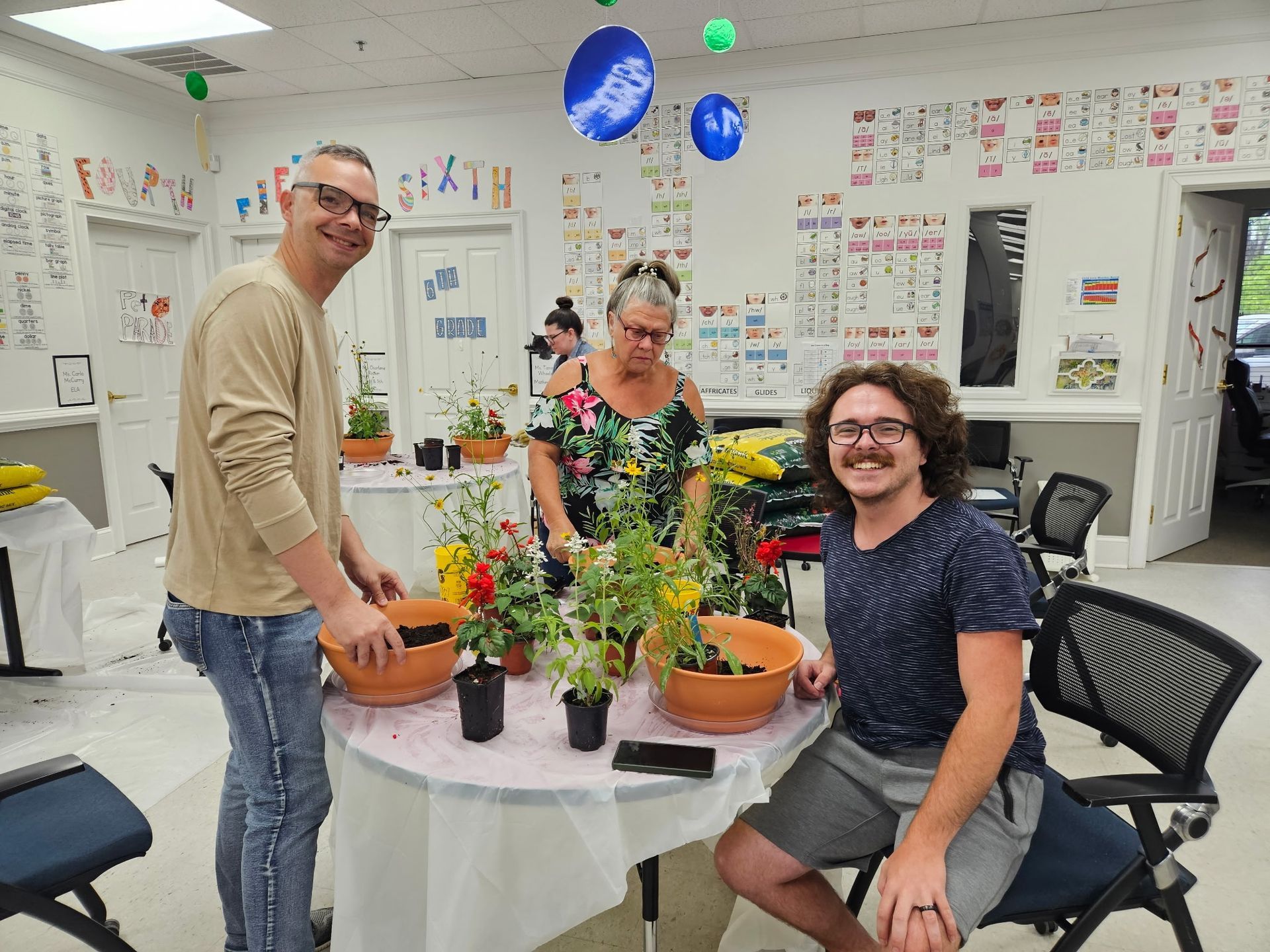 Three people smiling around a table with potted plants in a bright classroom or workshop