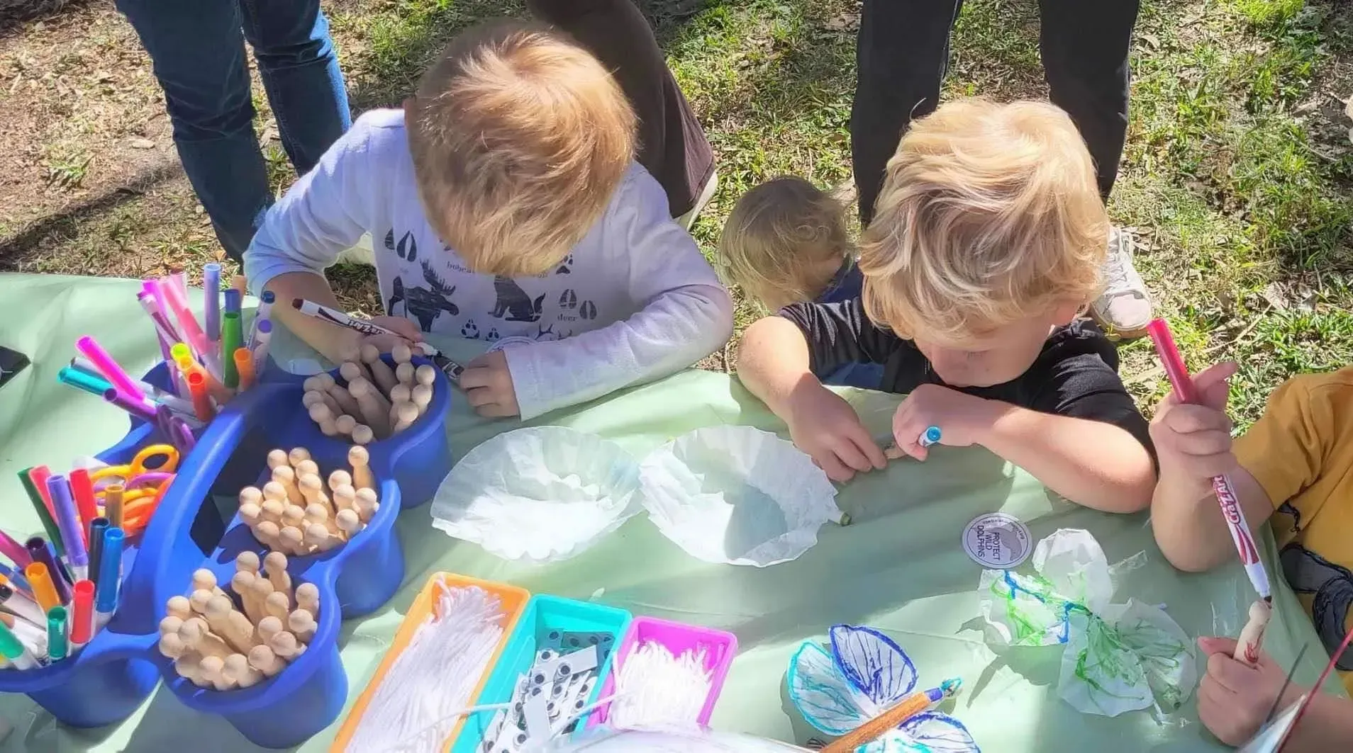 Children outdoors painting at a low table with colorful art supplies and craft materials