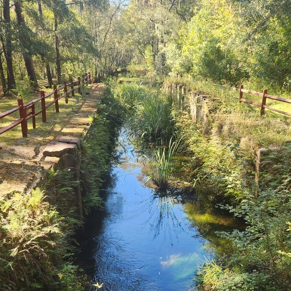 A narrow canal reflecting blue sky, lined with green trees and a dirt path with red railings on both sides.