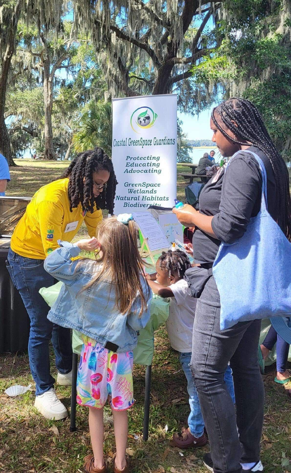People gathered outdoors near a sign, talking with a child in a colorful dress.