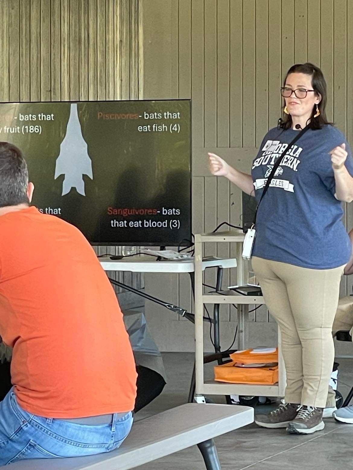 Person presenting beside a screen in a classroom, with another person seated in the foreground.