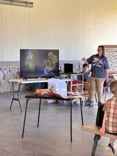 Person speaking into a microphone beside a TV display at a small indoor event.