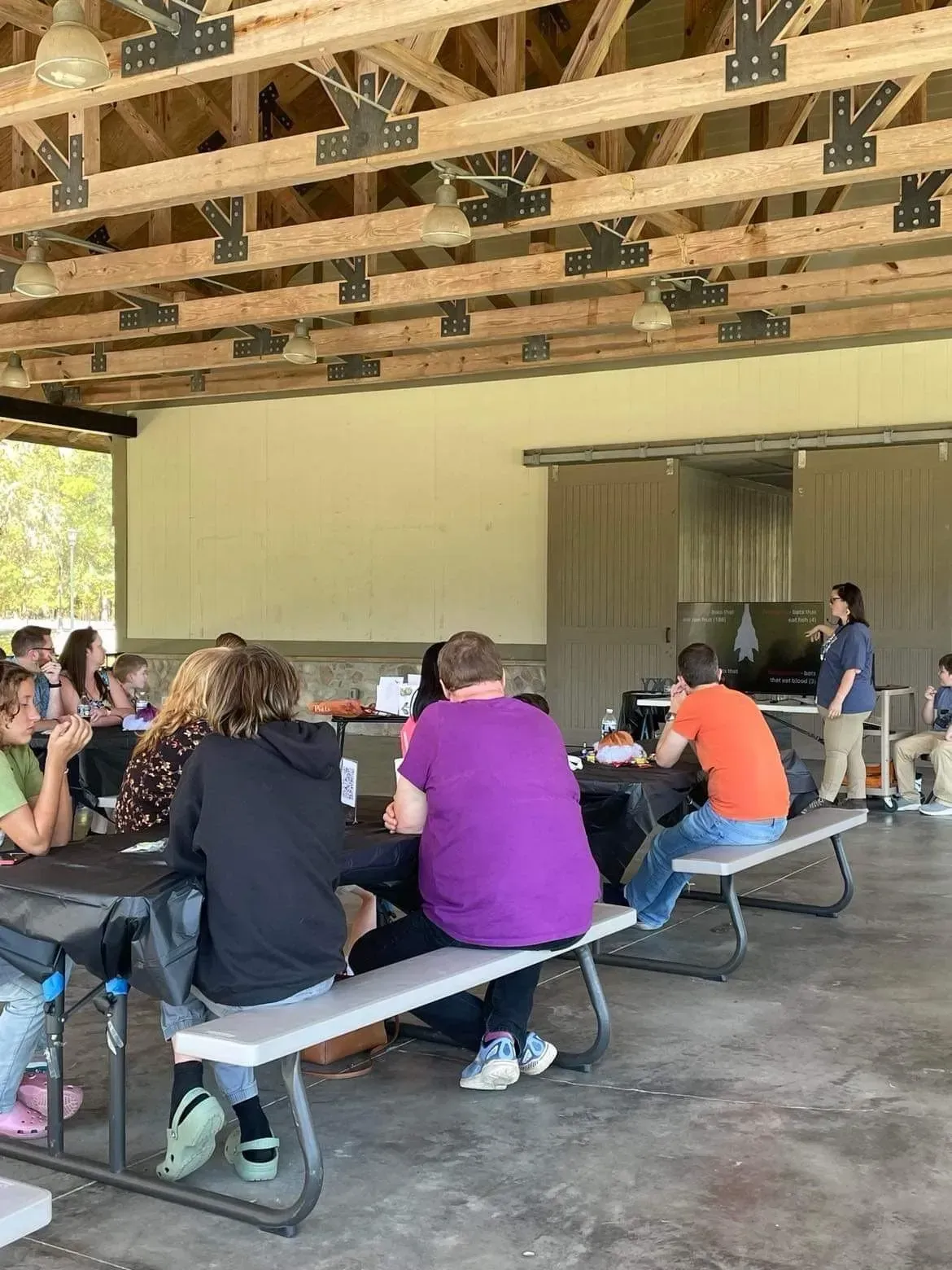 People seated at picnic tables in a covered pavilion, listening to a speaker near the front.