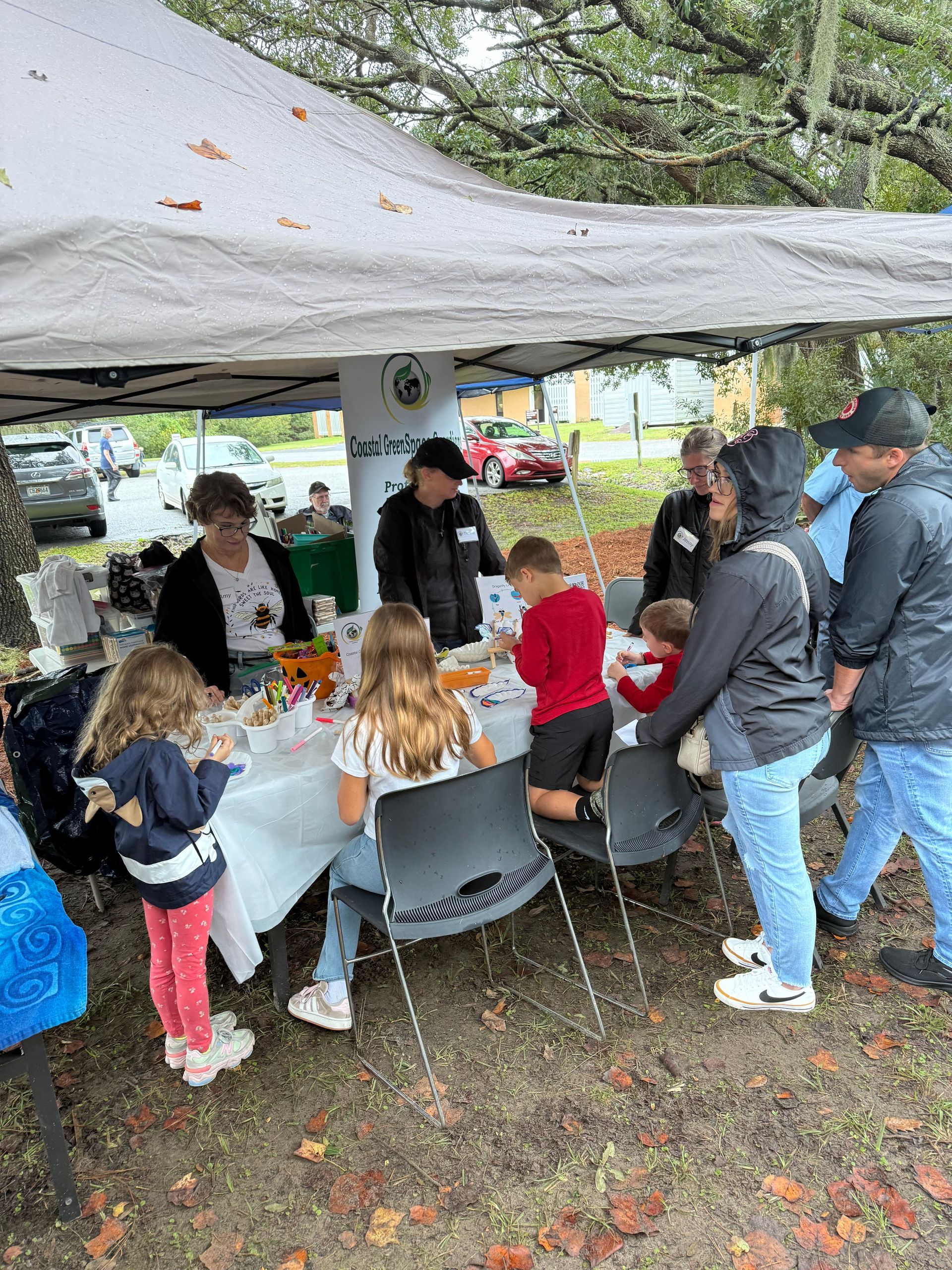 People gathered under a canopy at an outdoor table, serving food and chatting in a rainy setting.