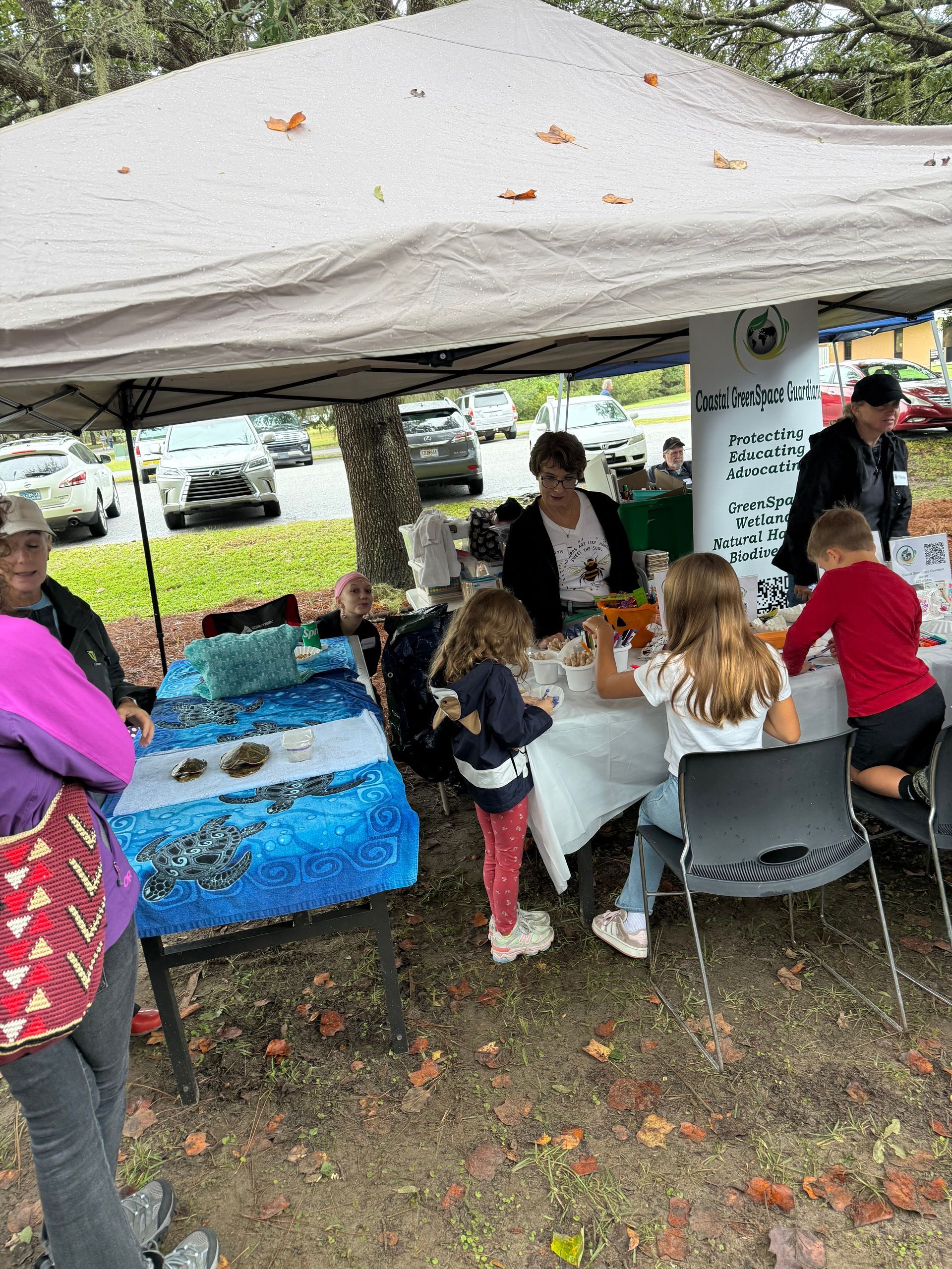 People gathered under a white canopy at an outdoor community booth with tables and chairs.