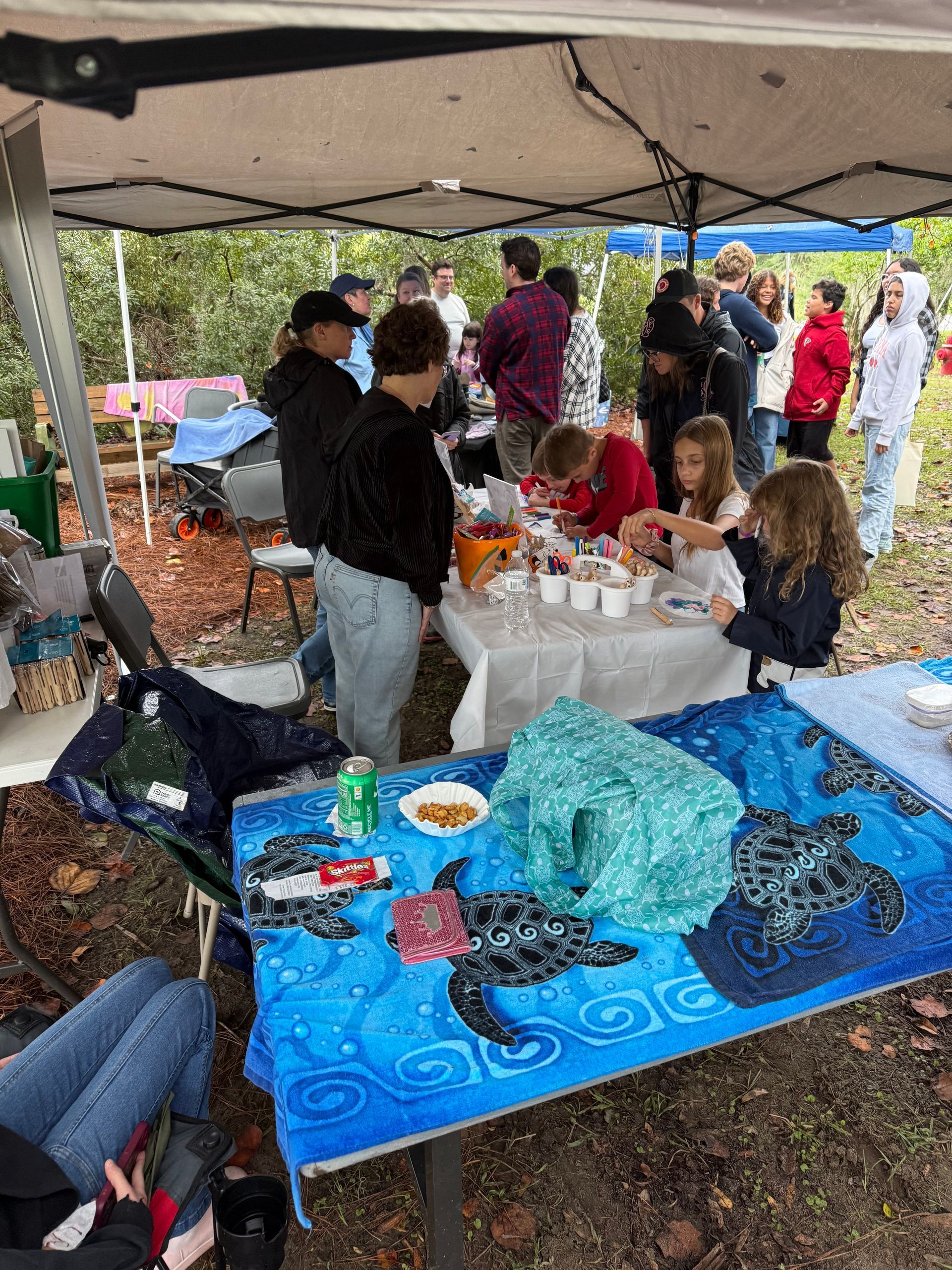 People gathered under a canopy at a picnic table with food and drinks, socializing outdoors.