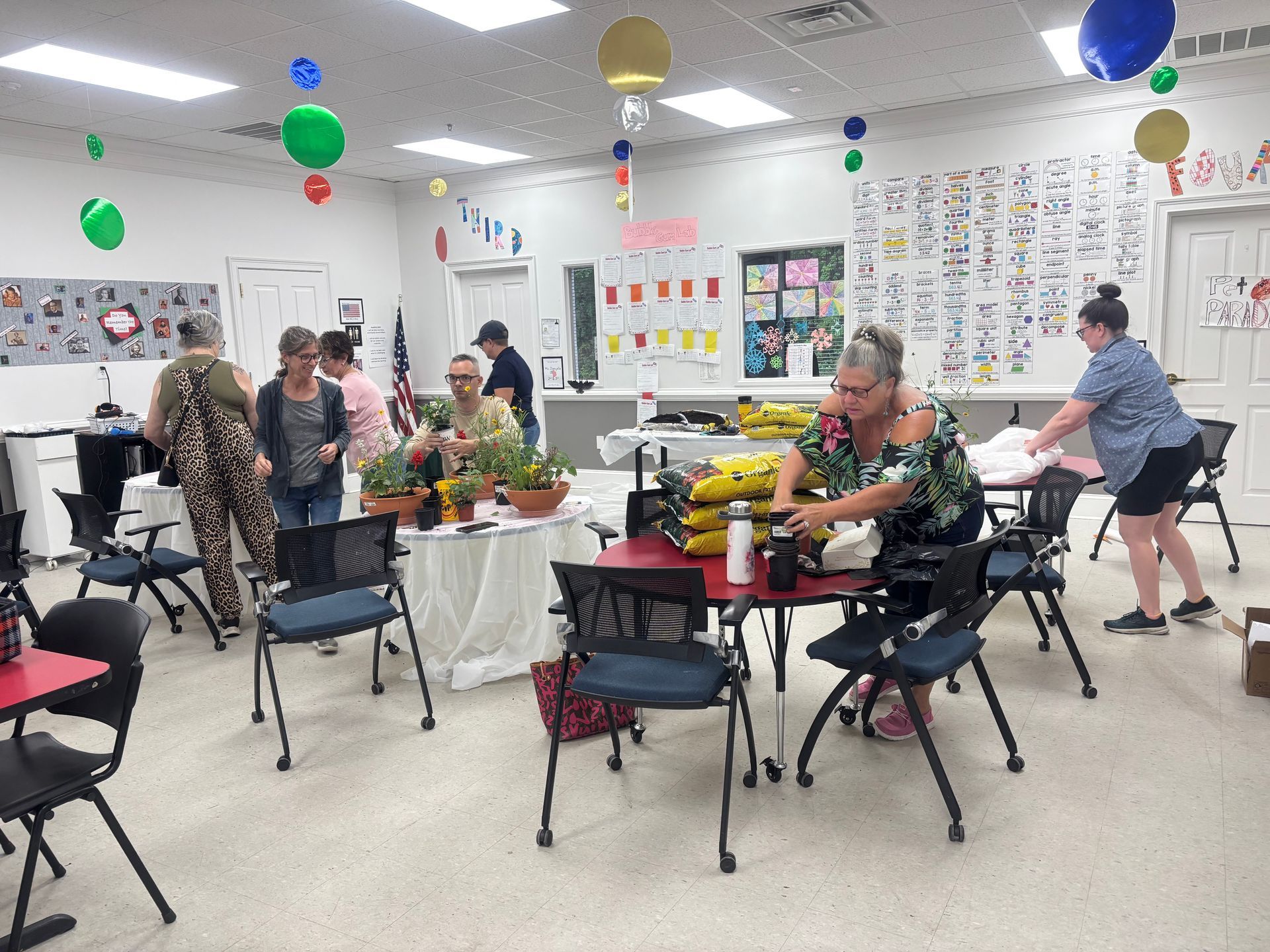 People gathered around tables in a bright room with balloons and posters, working on crafts and chatting.