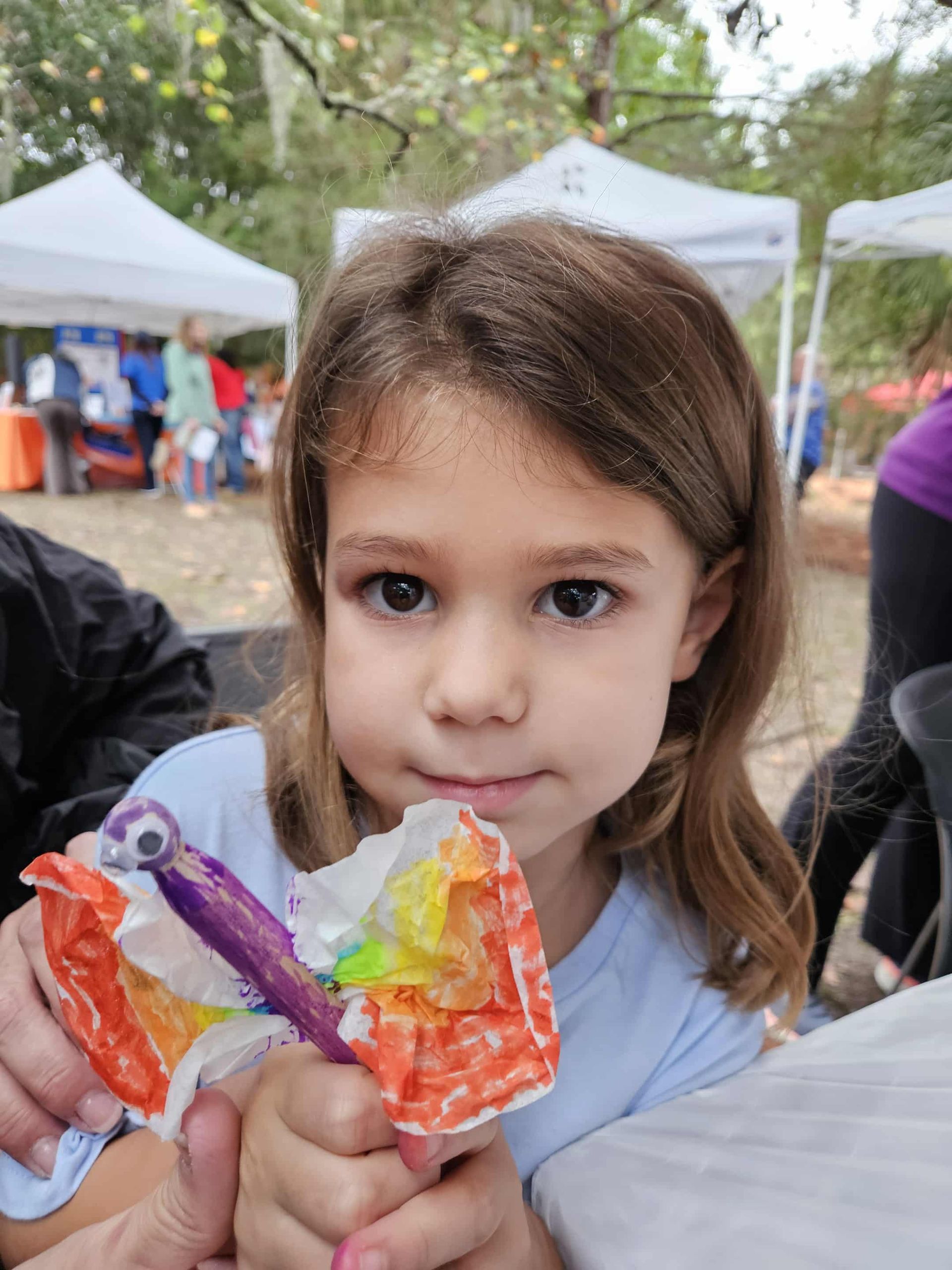 Child holding a colorful lollipop at an outdoor fair with white tents in the background