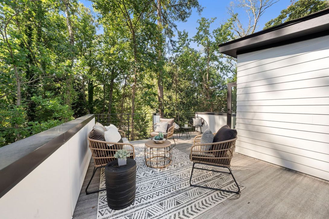 Outdoor deck with wicker chairs and table on a patterned rug beside a white house and trees.