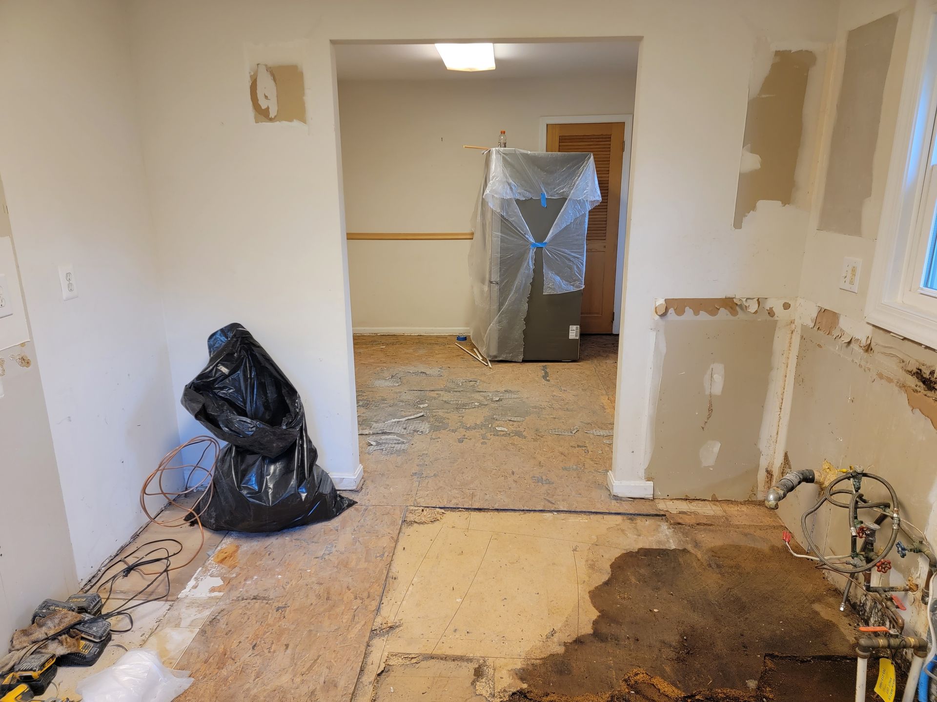 Partially renovated room with exposed floor, drywall damage, and a wrapped appliance in the back.