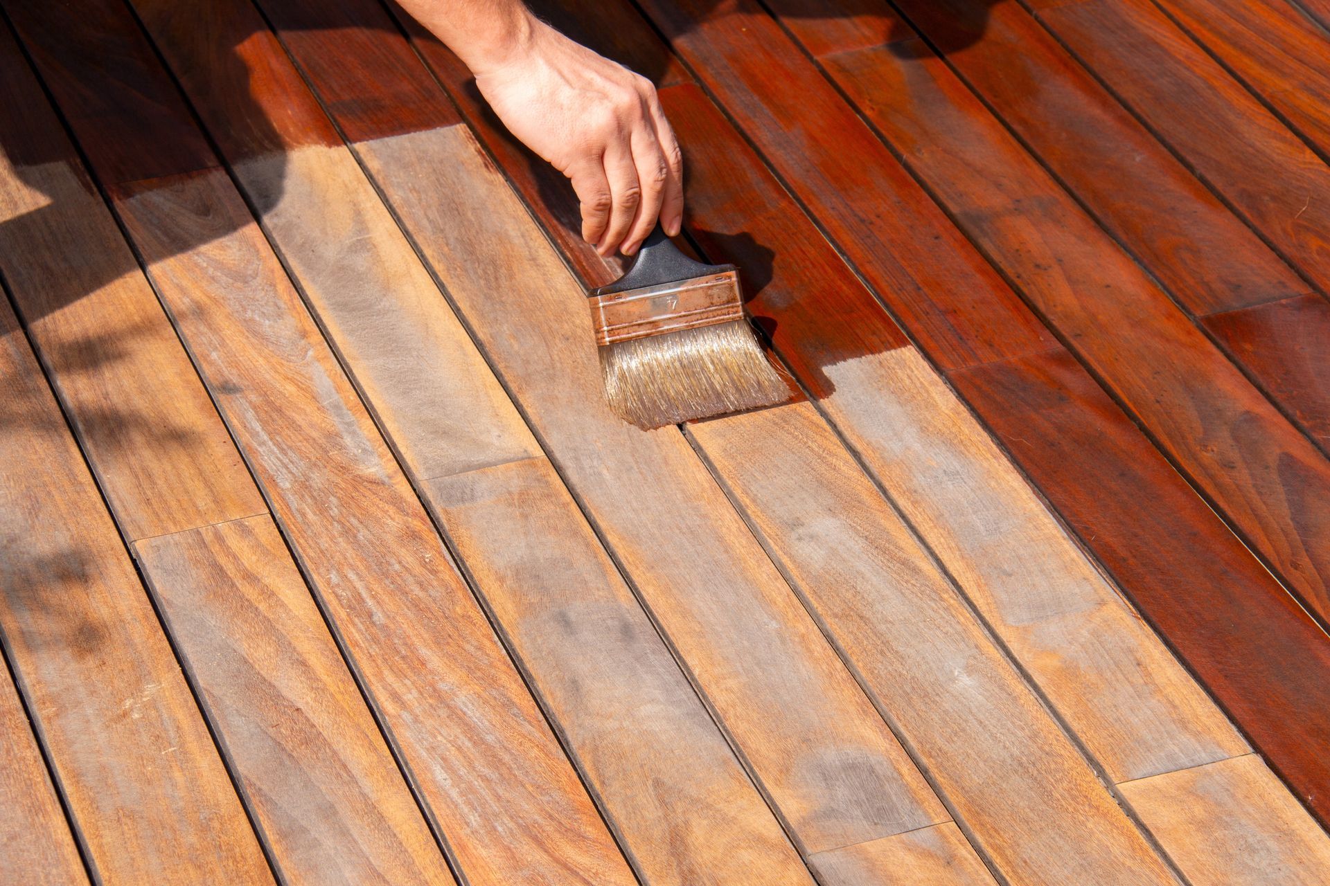A hand painting wooden deck boards with a brush, showing wet orange-brown stain and dry planks.
