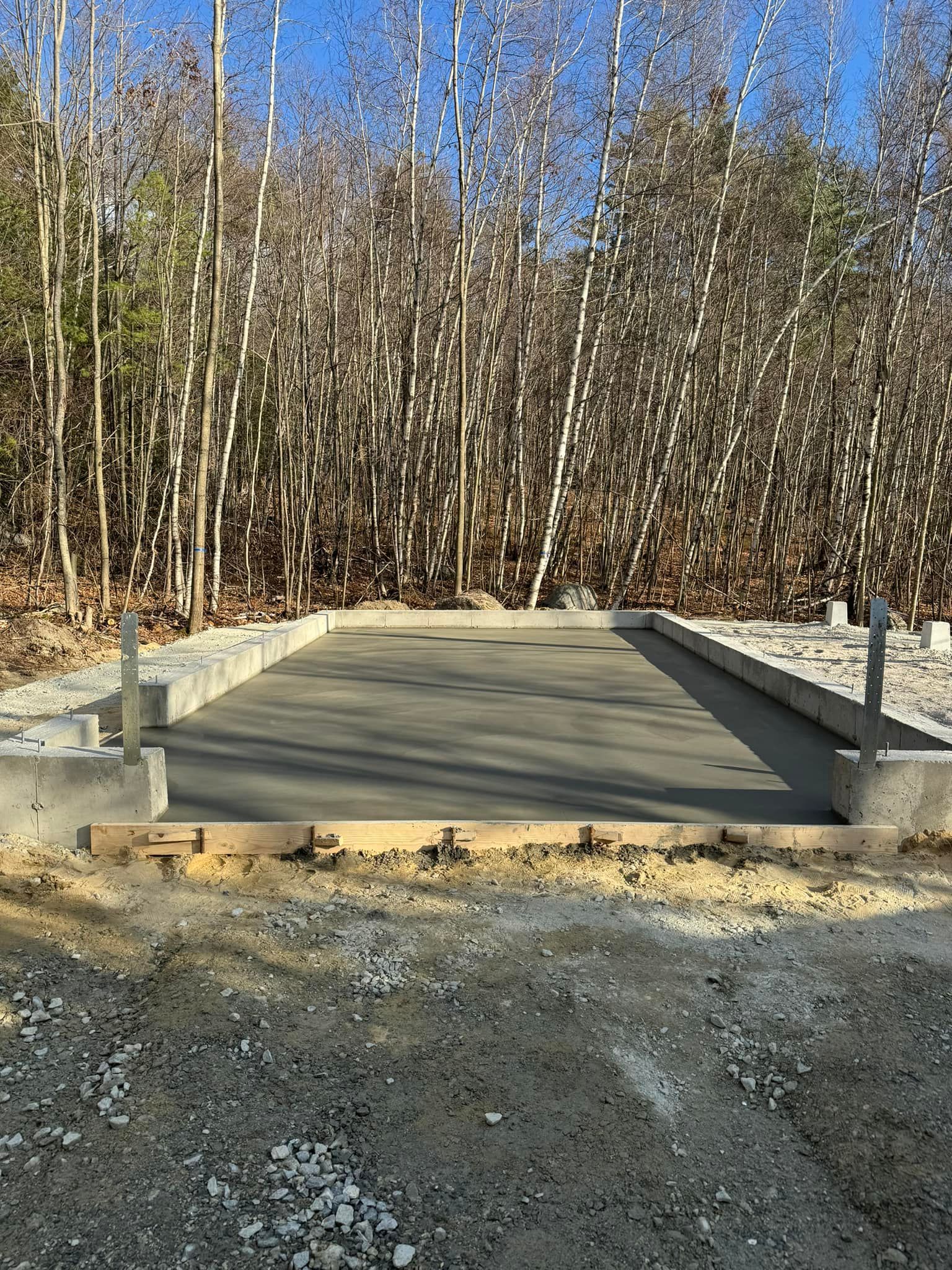 Concrete foundation, freshly poured, with wooden formwork and surrounding gravel, against a backdrop of bare trees under a clear blue sky.