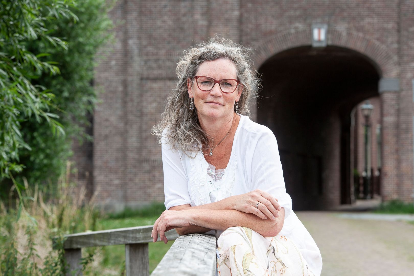 Vrouw met bril, leunend tegen een houten reling, glimlachend. Bij een bakstenen boogingang met begroeiing.