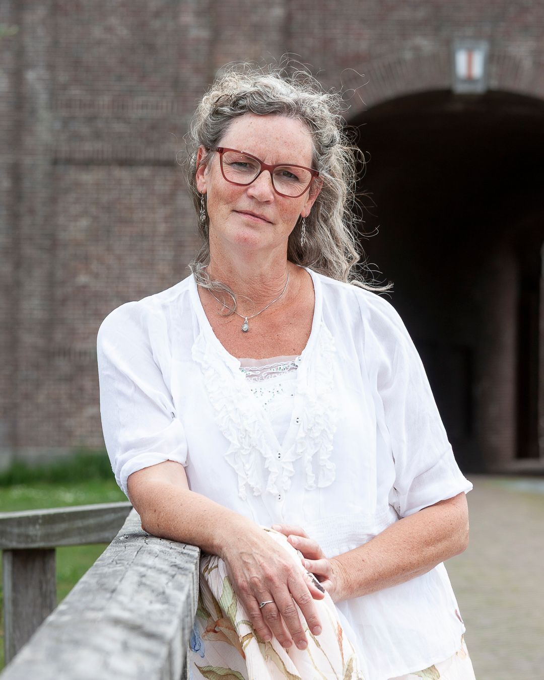 Een vrouw met een bril leunt tegen een houten reling, gekleed in een witte top, met een bakstenen gebouw op de achtergrond.