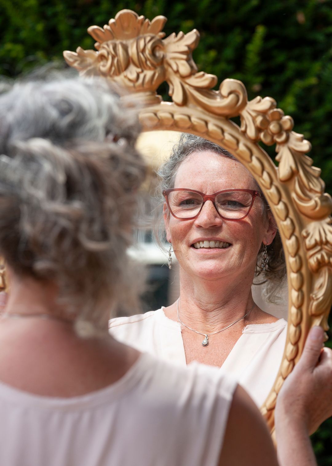 Vrouw glimlacht terwijl ze in een decoratieve gouden spiegel buiten kijkt.