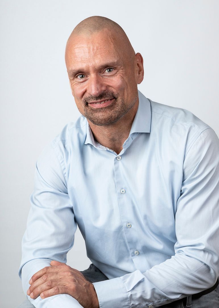 Een man met een kaalgeschoren hoofd, glimlacht en leunt, tegen een lichte achtergrond.