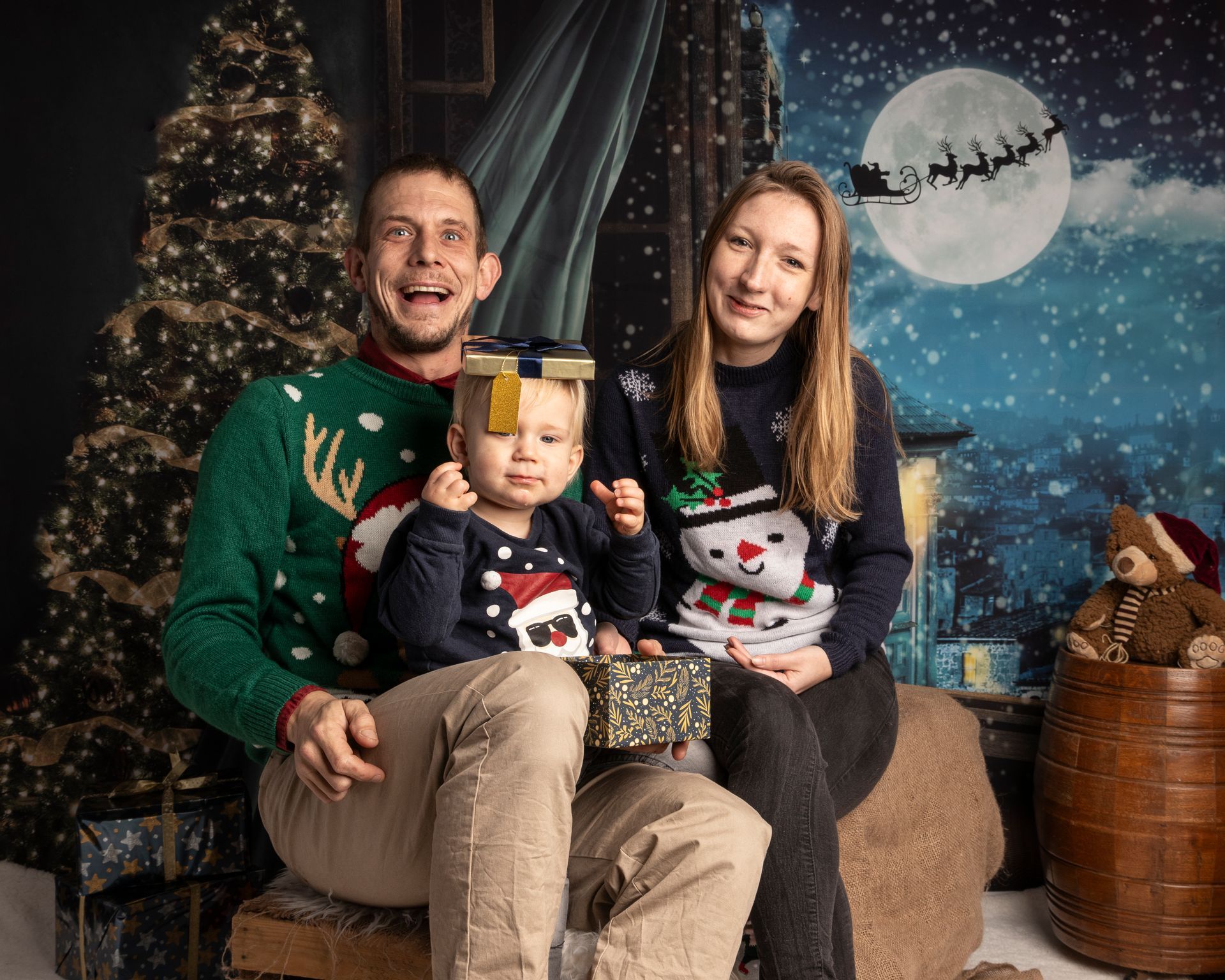 Een gezin in kersttruien poseert voor een foto tegen een feestelijke achtergrond. Een kind houdt een cadeautje vast.