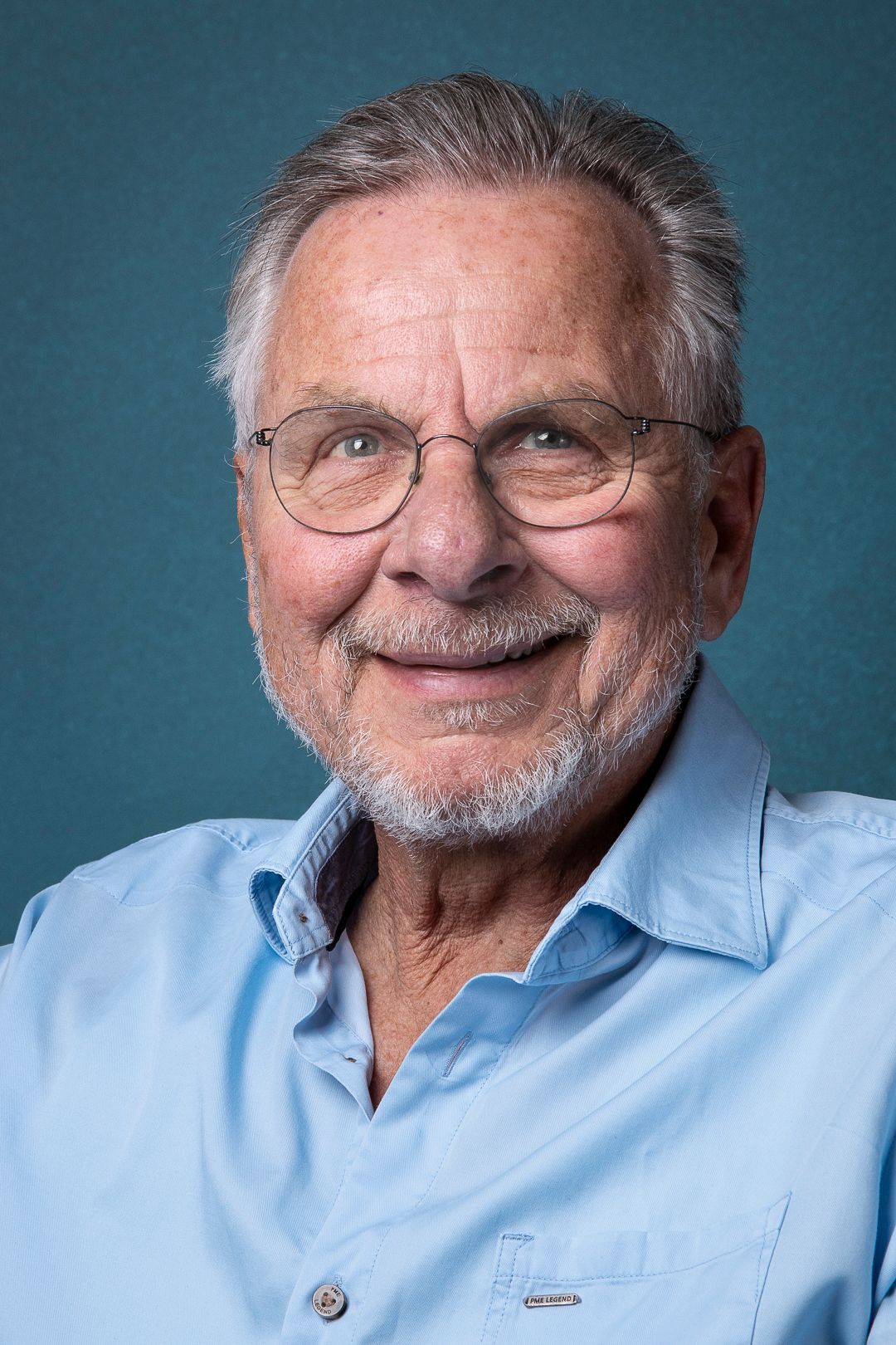 Lachende man met bril, lichtblauw shirt, tegen een turquoise achtergrond.
