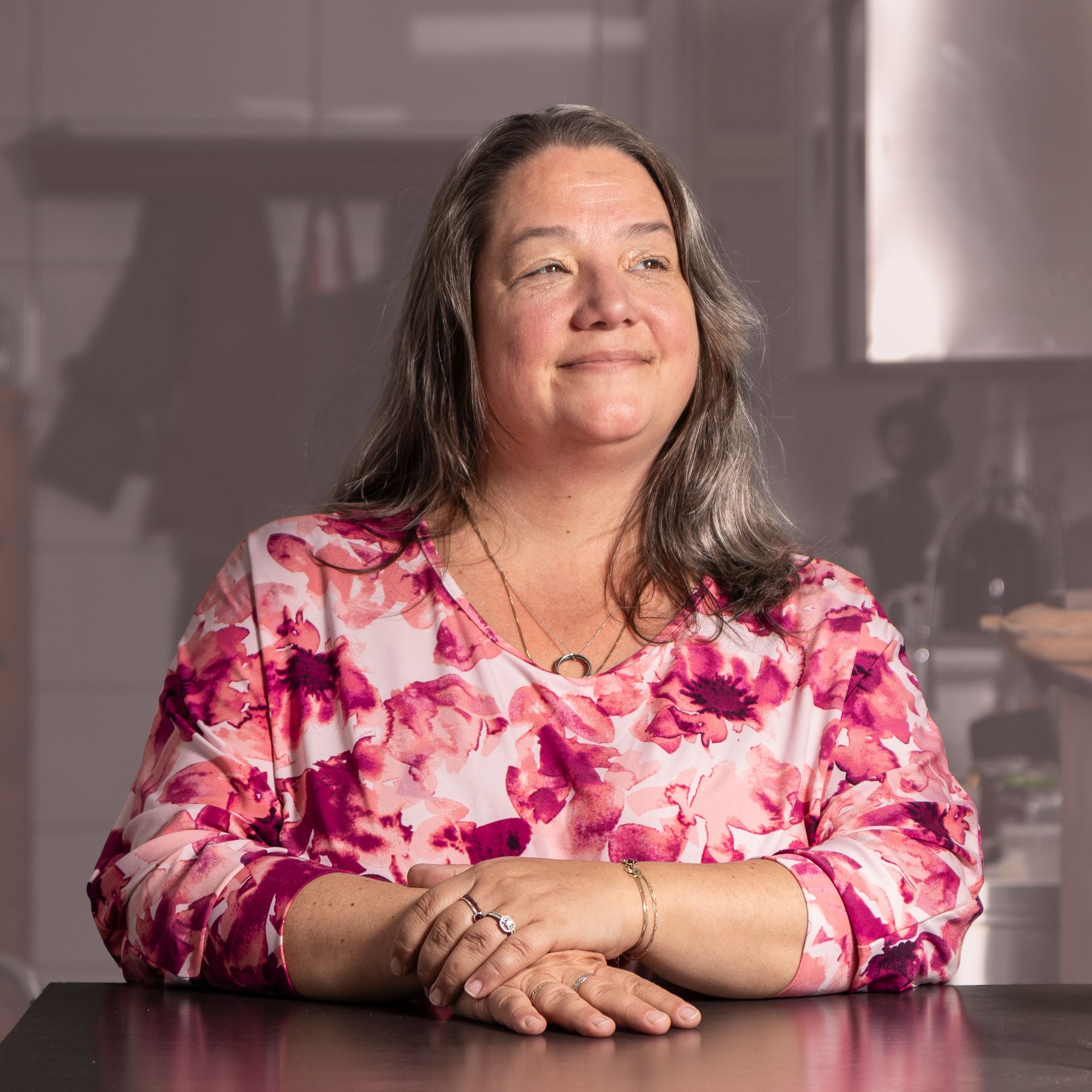 Vrouw in bloemenblouse, lachend met haar handen gevouwen op een tafel, keuken op de achtergrond.