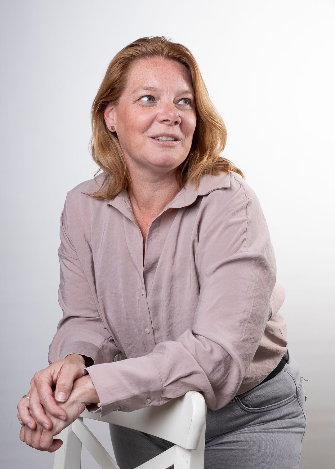Een vrouw in een mauve blouse leunt tegen een witte stoel en kijkt opzij, tegen een witte achtergrond.