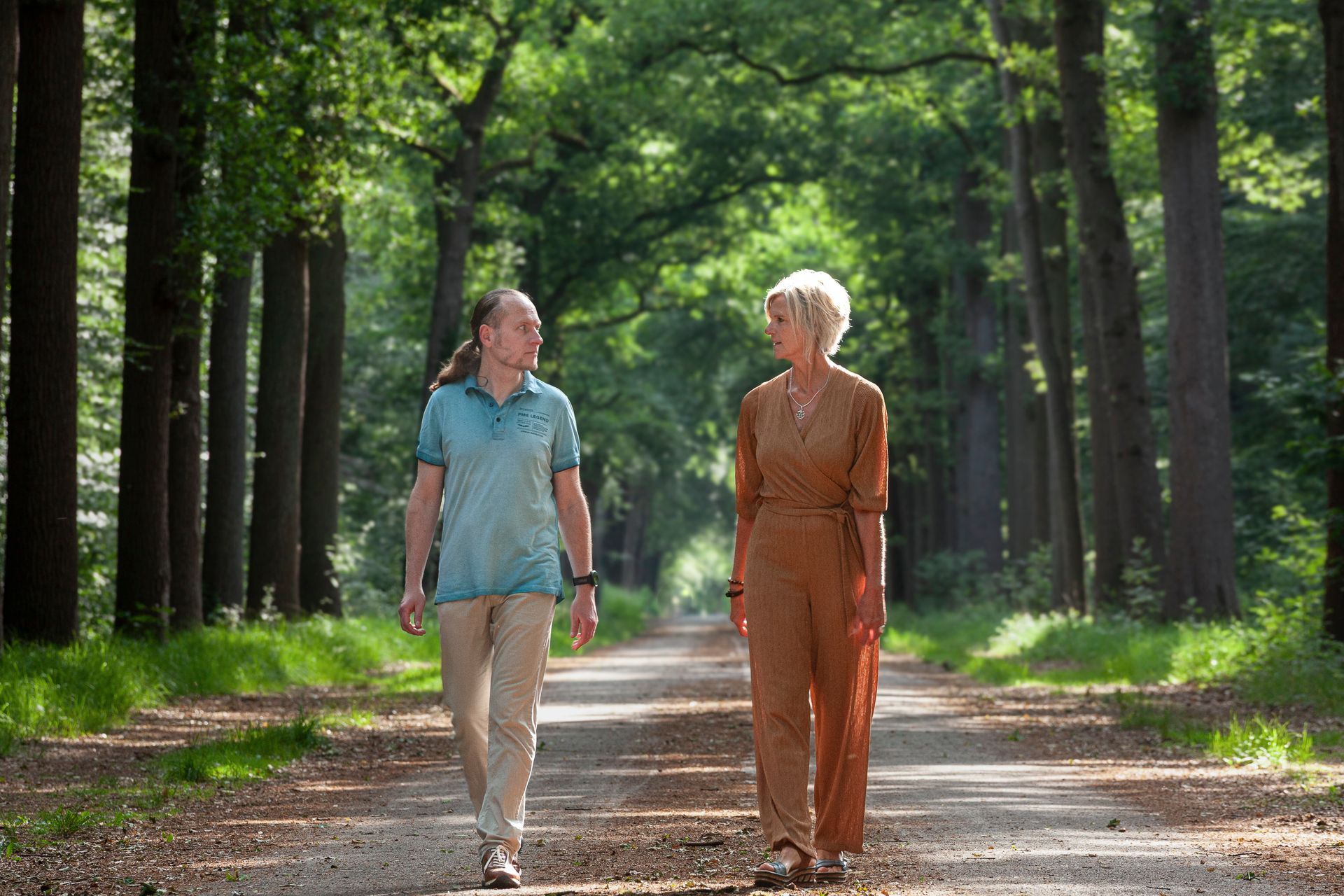 Twee mensen lopen over een pad door een zonovergoten bos. Ze zijn in gesprek met elkaar