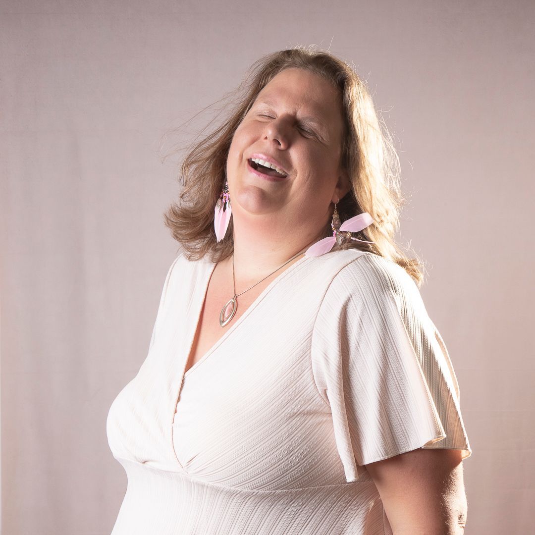 Vrouw in witte jurk lacht, ogen gesloten, roze oorbellen, achtergrond met zacht licht.