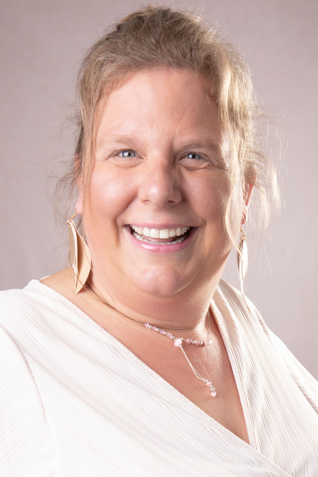 Een vrouw met blond haar lacht naar de camera, ze draagt een witte top en sieraden.