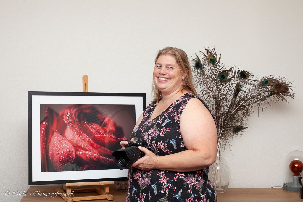 Een vrouw glimlacht, met een camera in haar hand, naast een ingelijste foto van een rode roos met waterdruppels.