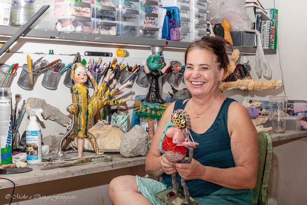 Vrouw zit aan een werktafel en houdt een pop vast. Werkplaatsomgeving, gereedschap en creaties zichtbaar.