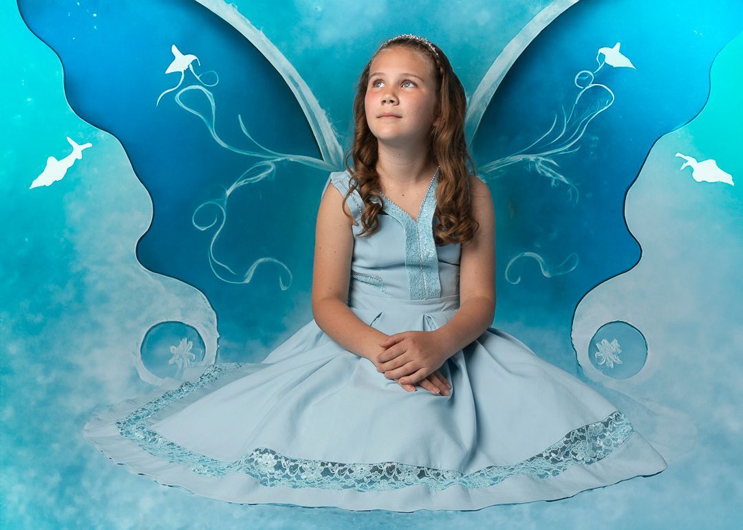 Meisje in een blauwe jurk met vlindervleugels, kijkt omhoog tegen een blauwe achtergrond.