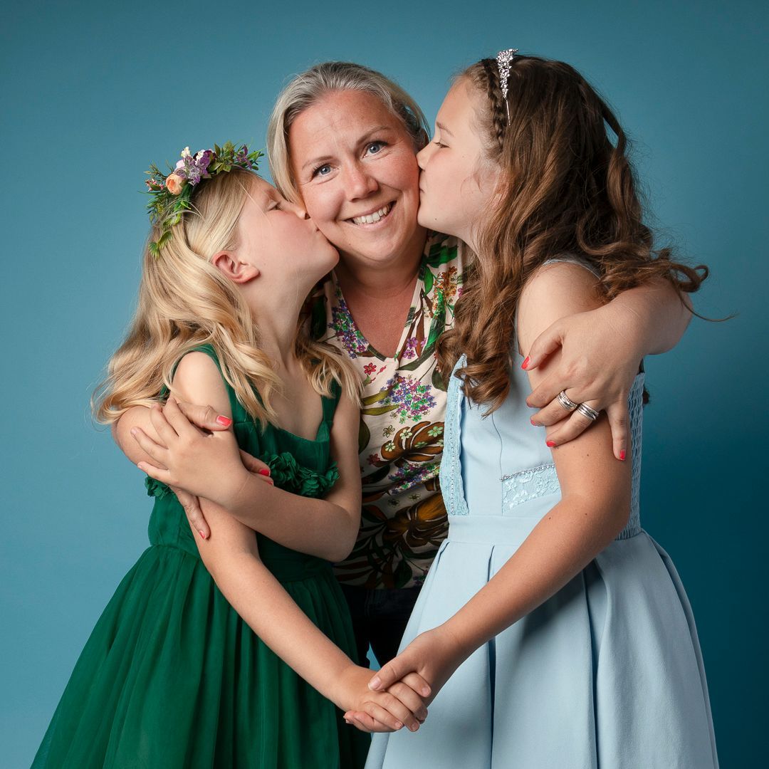 Vrouw omhelsd door twee meisjes, die kusjes op de wangen krijgt; blauwe achtergrond.