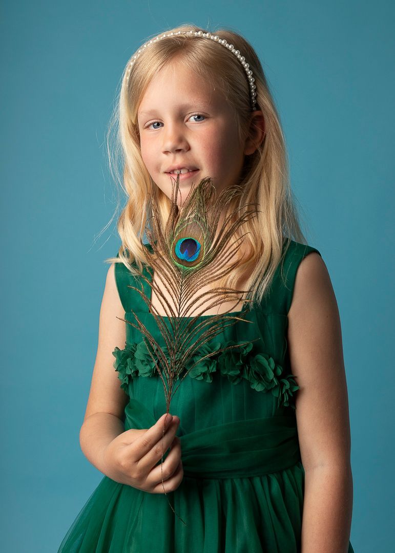 Een jong persoon in een groene jurk houdt een pauwenveer vast voor een blauwe achtergrond.