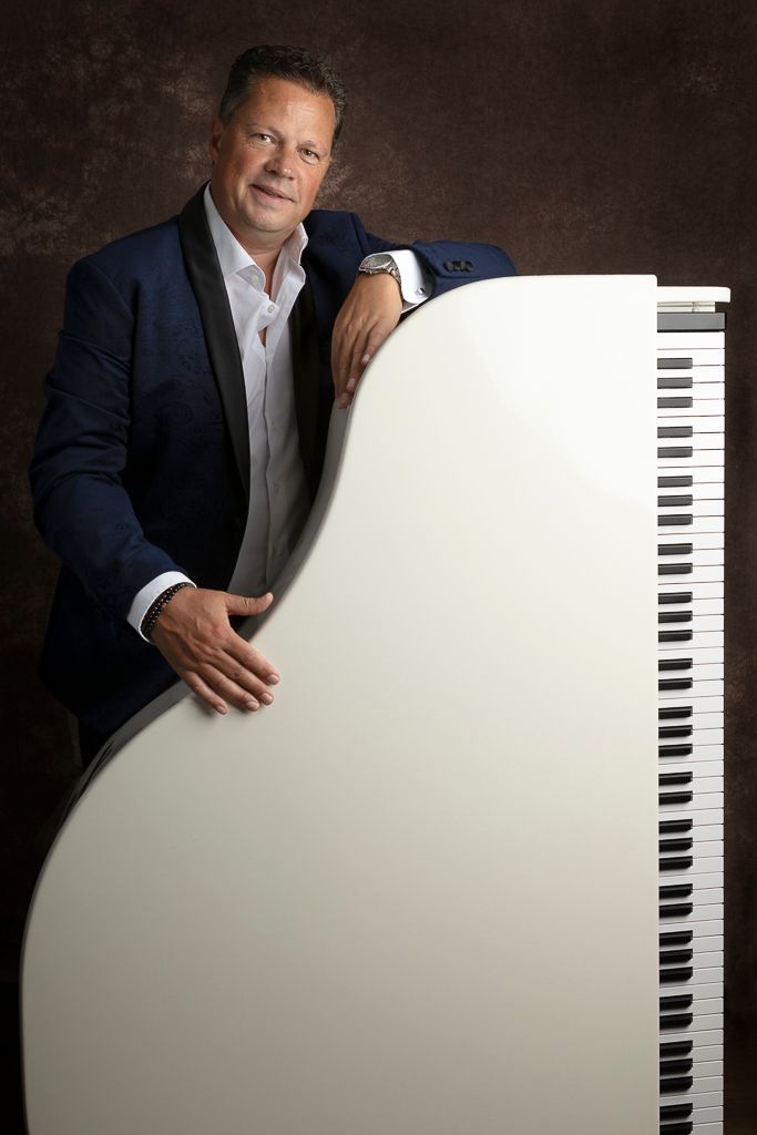 Een man in een blauwe blazer leunt tegen een witte piano. Bruine achtergrond.