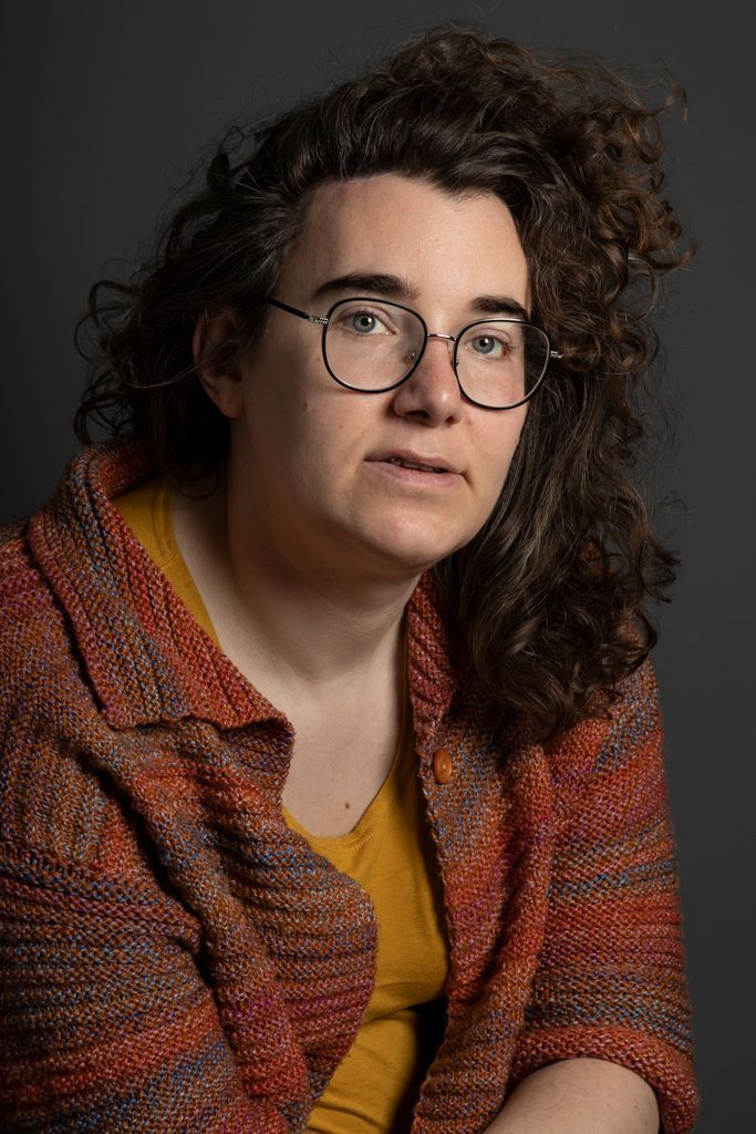 Vrouw met krullend haar en een bril, kijkt voor zich uit tegen een grijze achtergrond.