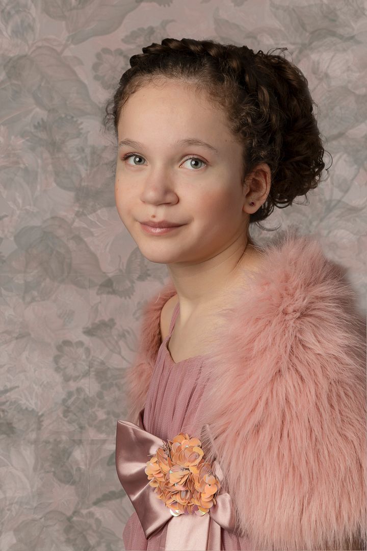 Meisje in roze jurk en bontstola, lachend, met gevlochten opgestoken haar, bloemenbroche op een grijs patroonachtergrond.