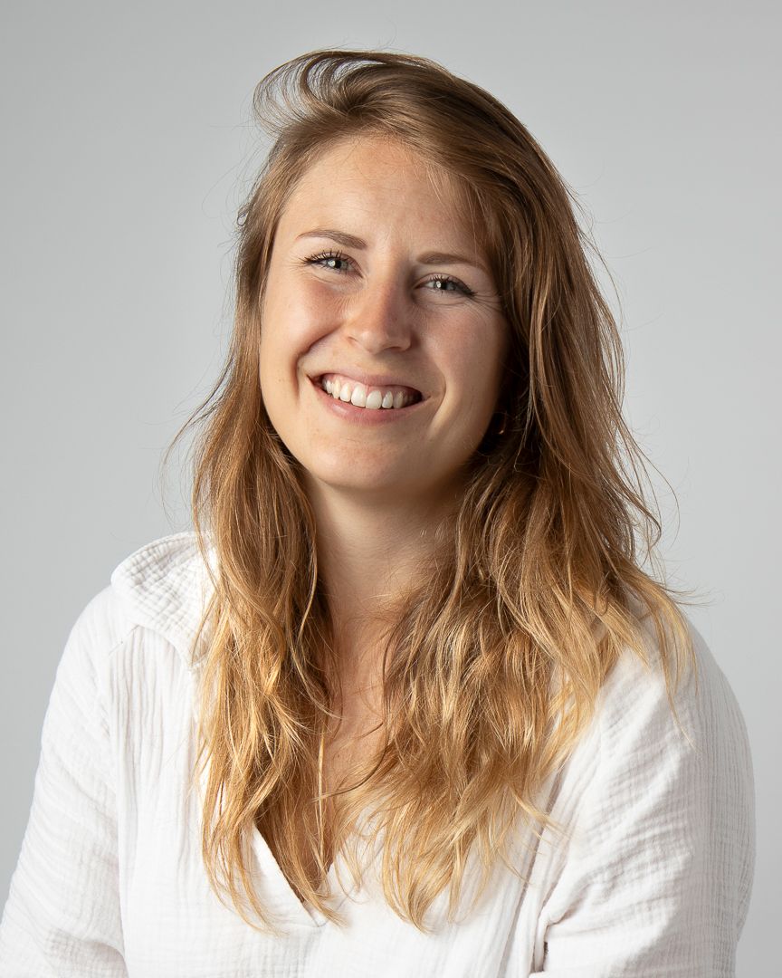 Vrouw met blond haar lacht, draagt een witte blouse en staat voor een neutrale achtergrond.