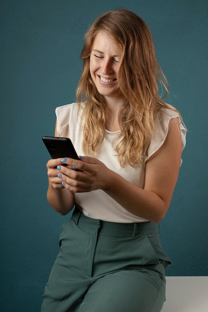 Een vrouw glimlacht naar haar telefoon, gekleed in een witte top en een groene broek, en leunt tegen een turquoise muur.