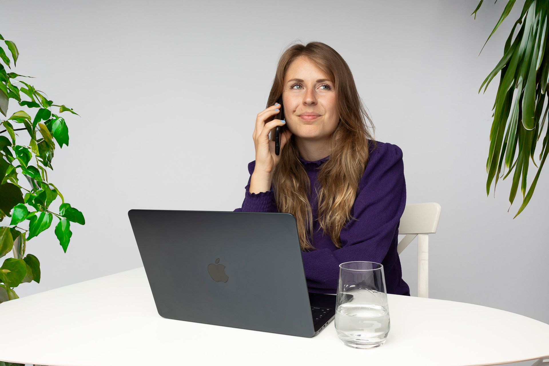 Vrouw aan de telefoon, aan het werk voor een laptop, zittend aan een witte tafel met planten.