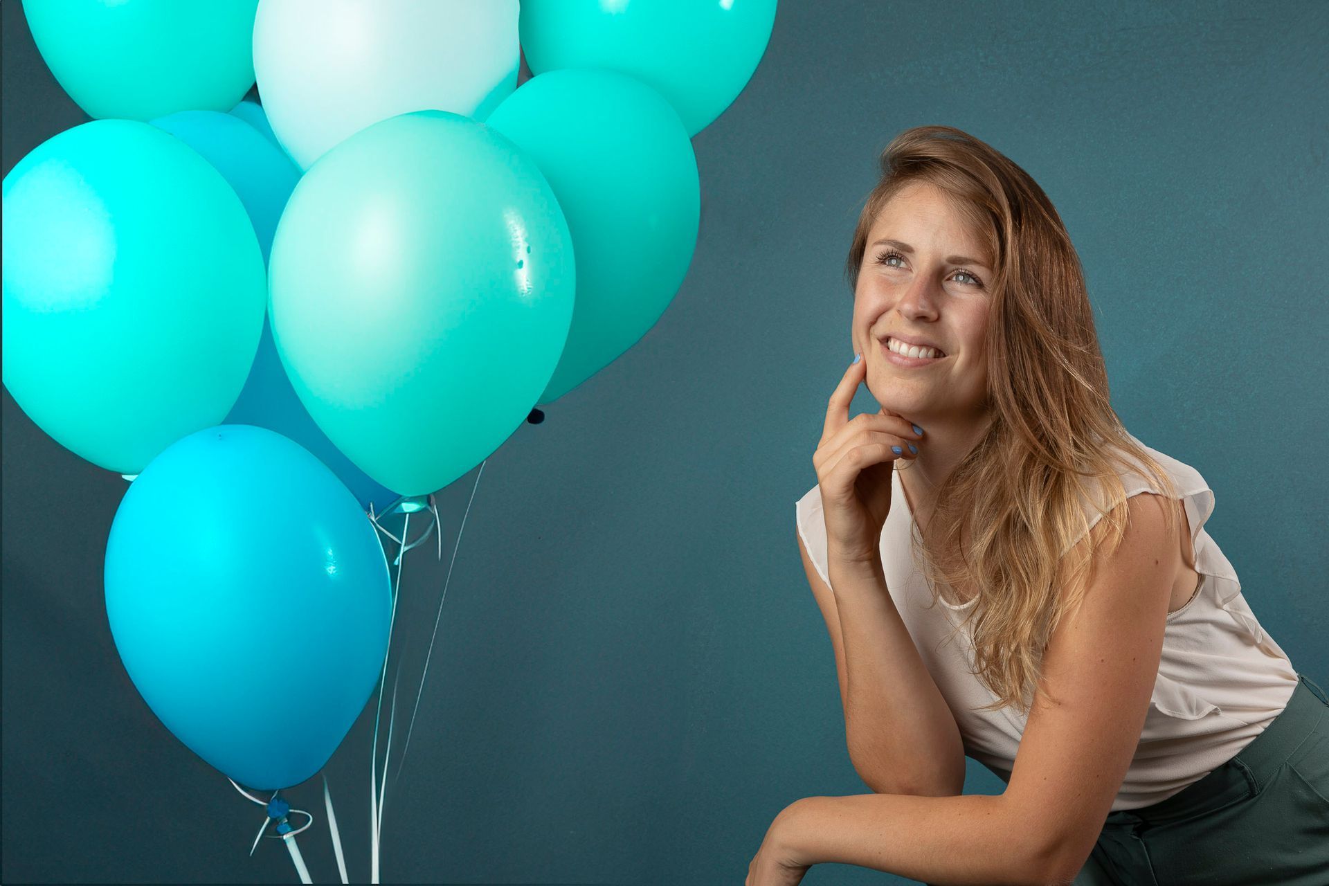 Een vrouw glimlacht en kijkt omhoog. Ze houdt ballonnen vast en leunt tegen een blauwe achtergrond.