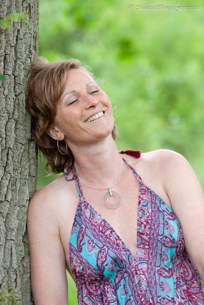 Een vrouw leunt tegen een boom en glimlacht met gesloten ogen.  Buiten, groene achtergrond.