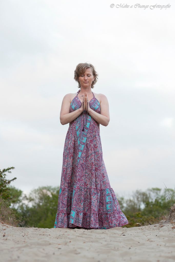 Vrouw in halterjurk, handen ineengevouwen, buiten op een pad. Lichtroze, blauwe en paarse jurk, bewolkte lucht.