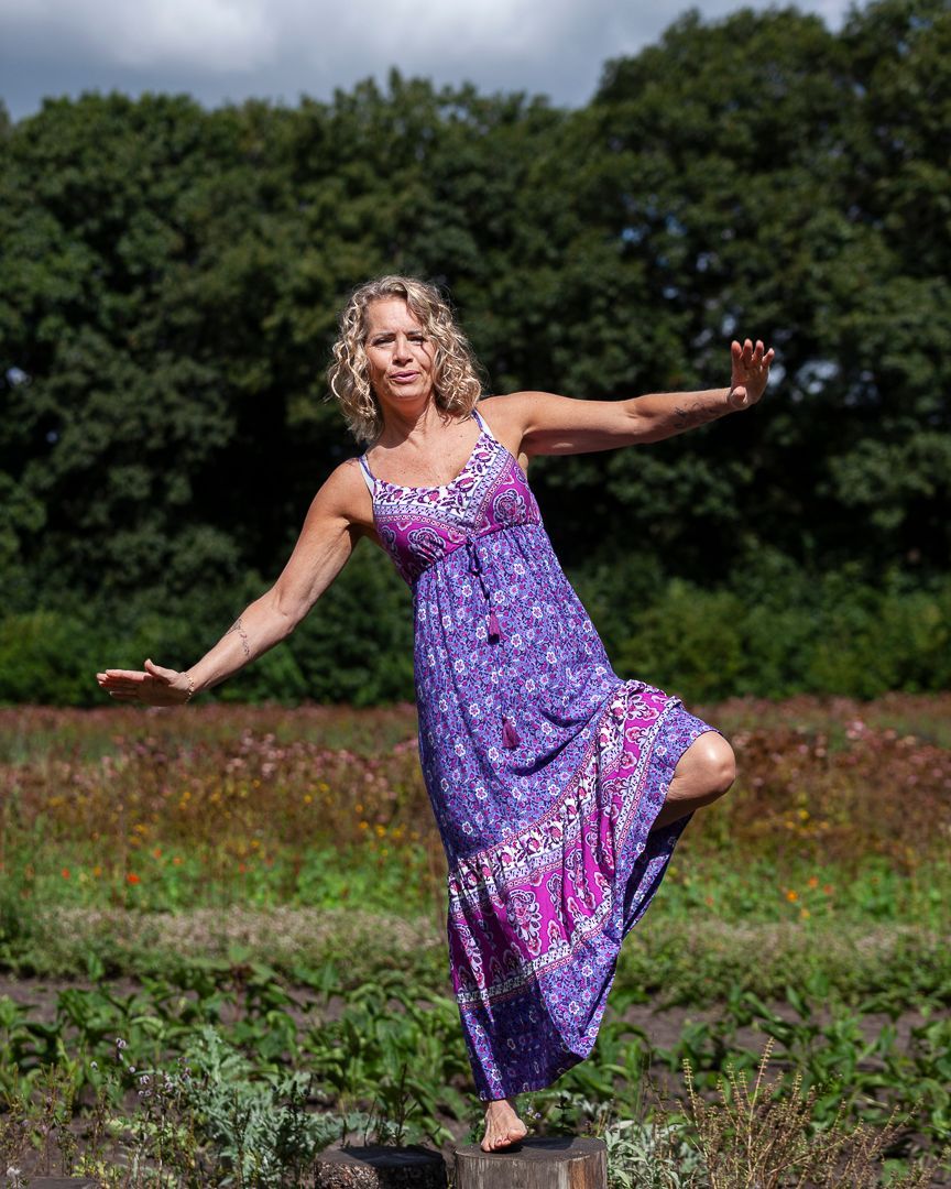 Een vrouw in een paarse jurk balanceert op één been, met uitgestrekte armen, in een veld met bomen.