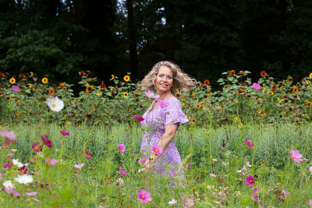 Een vrouw in een paarse jurk lacht in een bloemenveld. Zonnebloemen en roze cosmos staan in bloei.