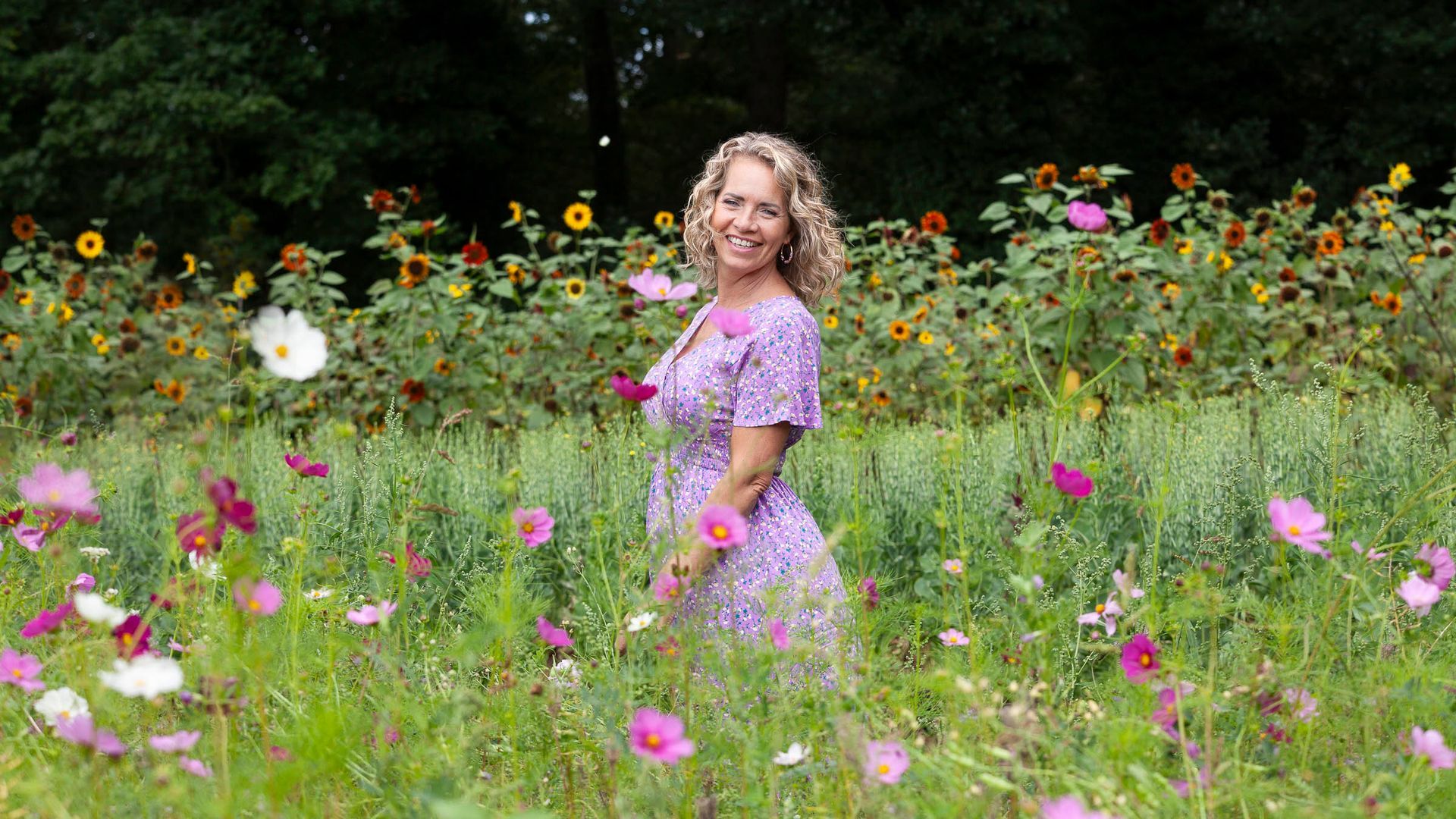 Een vrouw glimlacht te midden van een kleurrijk veld met wilde bloemen, met zonnebloemen op de achtergrond.
