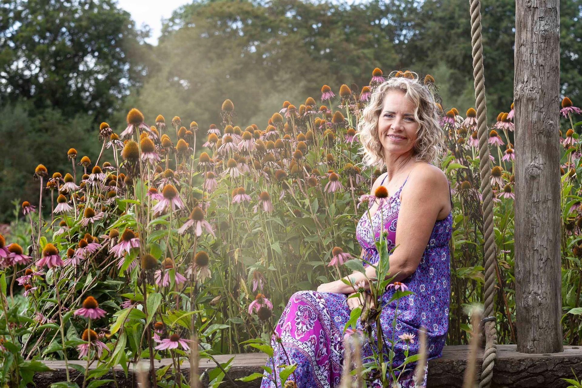 Een vrouw in een paarse jurk zit glimlachend op een schommel in een bloementuin.