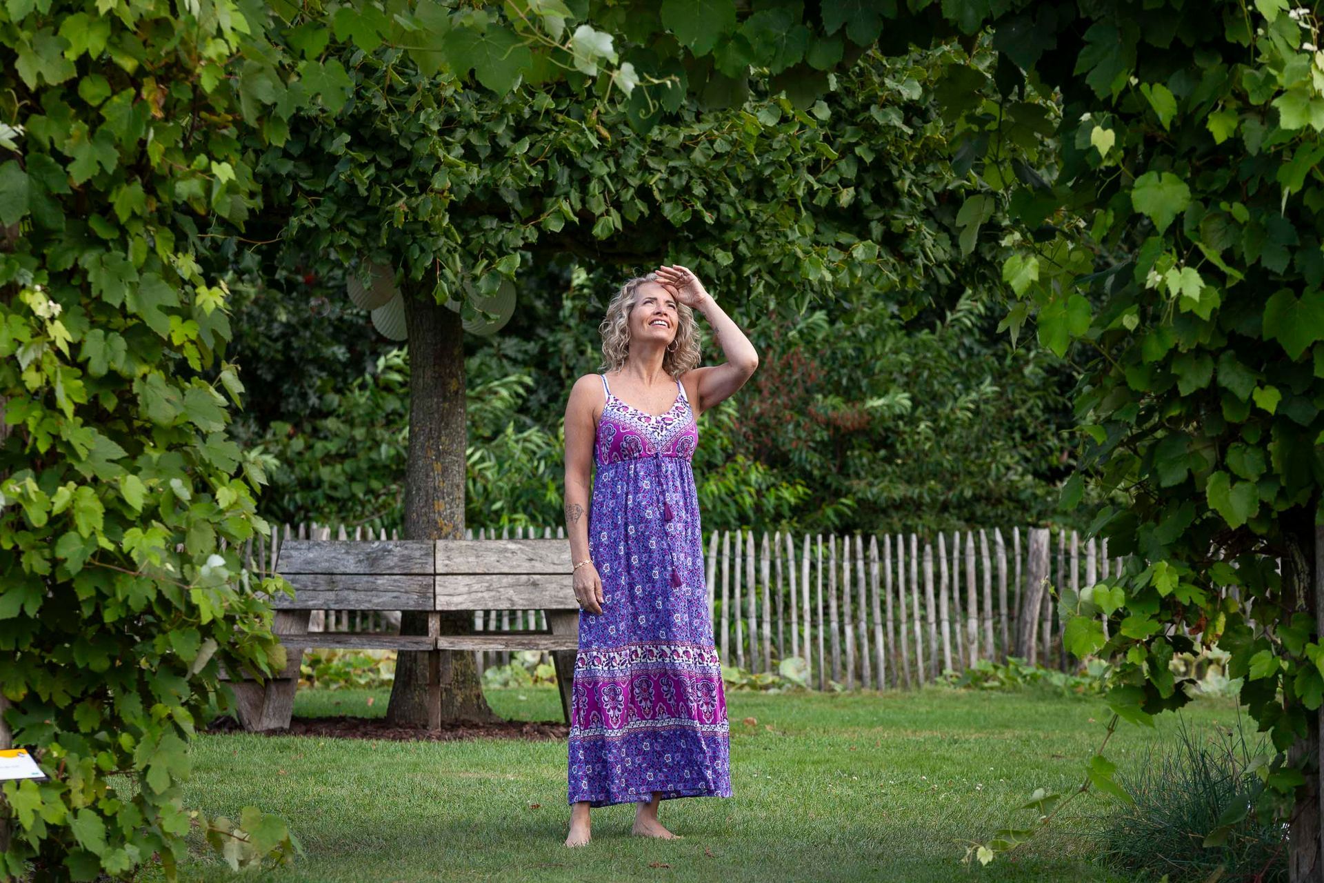 Vrouw in paarse jurk staat in een tuin, met haar armen omhoog naar de zon, onder een groene boog van klimplanten.