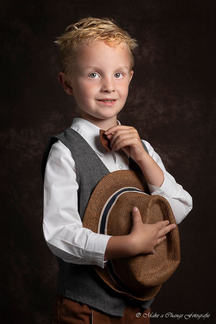 Jongen in vest en wit shirt houdt een hoed vast, lacht naar de camera, bruine achtergrond.