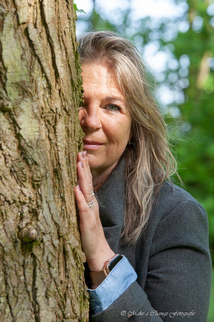 Vrouw gluurt achter een boom vandaan, hand op de stam, glimlachend, grijze jas aan, buiten.