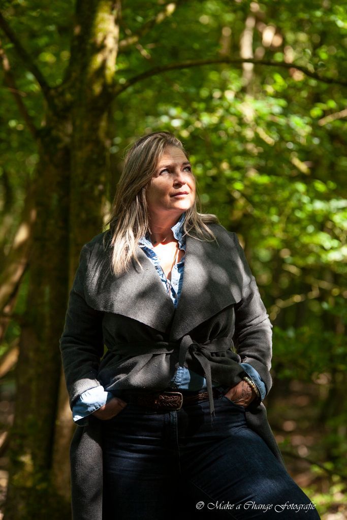 Vrouw in een bos, gekleed in een grijze jas, kijkt omhoog. Zonlicht schijnt door de bomen.