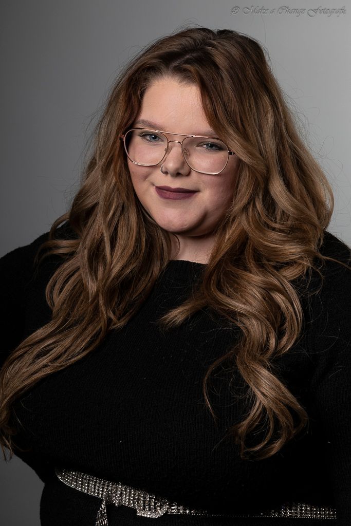 Vrouw met lang, golvend bruin haar, een bril en een zwarte trui aan, die lichtjes glimlacht.